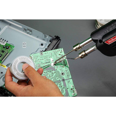 An action shot of the Weller professional dual-heat soldering gun being used to repair a circuit board, demonstrating the precision of the heavy-duty tip and the effectiveness of the integrated work lights.