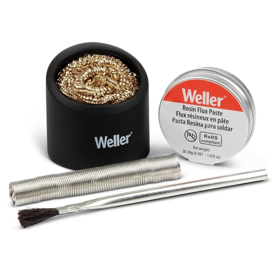 A studio product shot of the Weller WCACCK2 soldering accessory kit including lead-free solder wire, a tin of rosin flux paste, an application brush, and a brass wire tip cleaner in a black silicone holder.