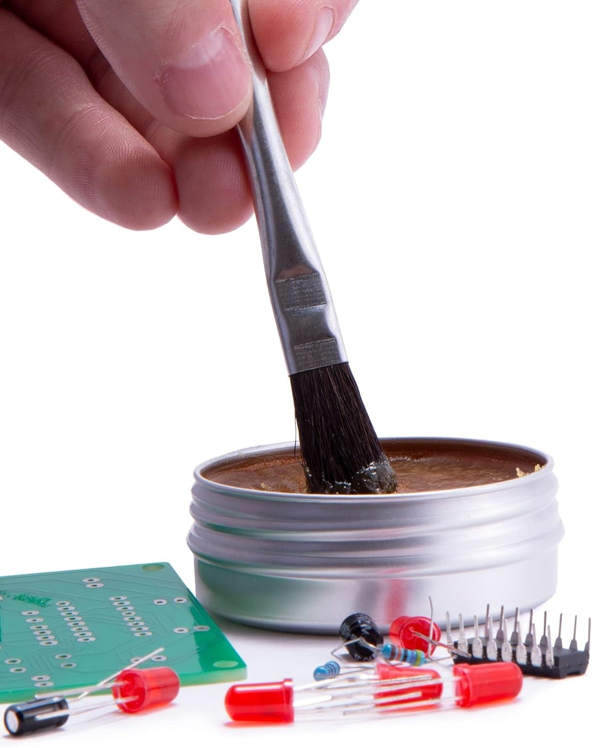 A detailed shot of a hand using the metal flux brush to apply professional-grade rosin flux paste from the Weller tin to an electronic circuit board for improved solder wetting and flow.