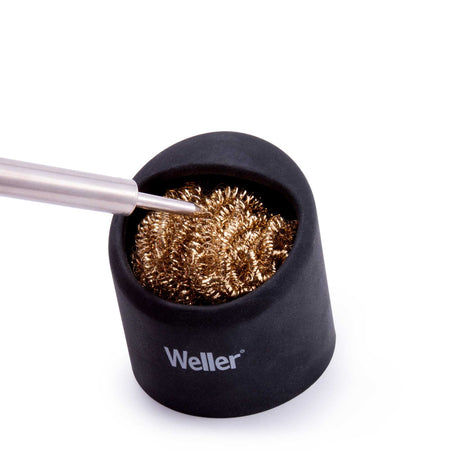 A professional soldering iron tip being plunged into a Weller brass sponge cleaner held in a black silicone stand to demonstrate the efficient waterless cleaning process.
