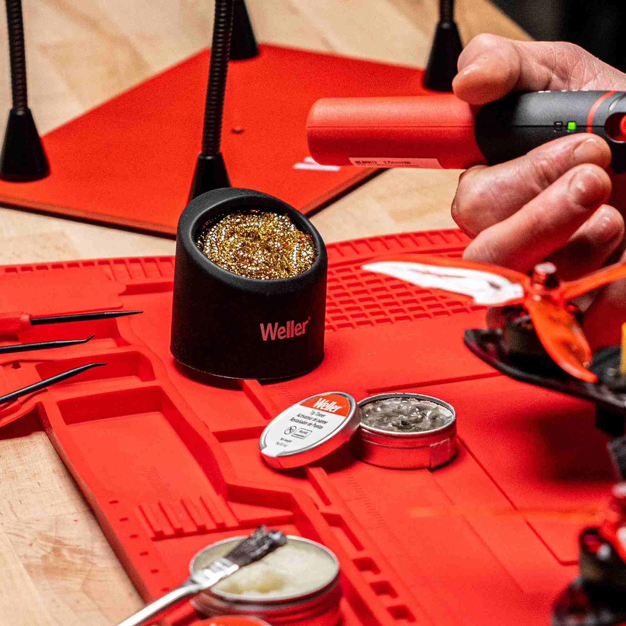 Action shot of a technician using a Weller brass sponge tip cleaner at an electronics repair workstation, surrounded by tools, a red heat-resistant mat, and a circuit board project.