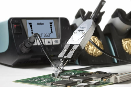 Close-up of a PCB being soldered with heated tweezers connected to the Weller WT2021MS station. The display shows 350°C. Smoke rises from the contact point, indicating active soldering. Brass wire sponges and tip holders are visible in the background.