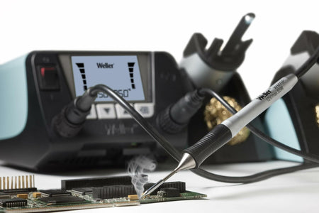 Live soldering scene featuring the Weller WT2021MS station set to 350°C. A Weller WXP 120 soldering iron is actively soldering a circuit board, with visible smoke rising from the contact point. Brass wire sponges and tip holders are shown in the background.