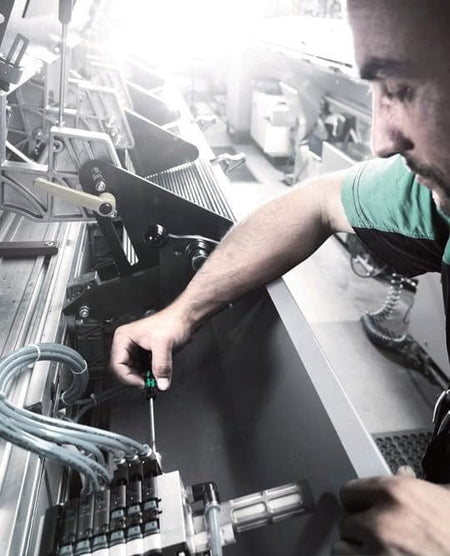 Person using a screwdriver to adjust industrial machinery with wires, connectors, and mechanical components in a workshop setting.