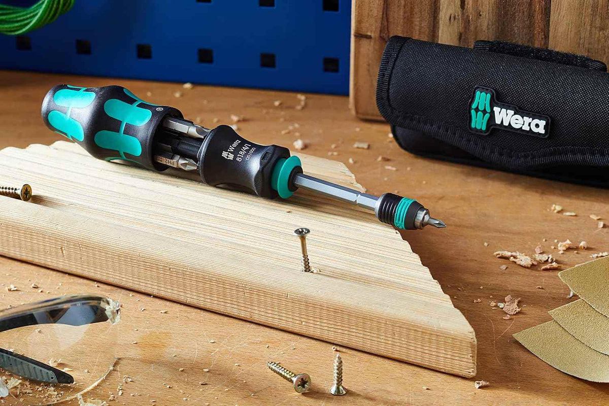 A lifestyle image of the Wera 05051021001 set on a wooden workbench, showcasing the screwdriver with the bit magazine open alongside the textile storage pouch and various wood screws.