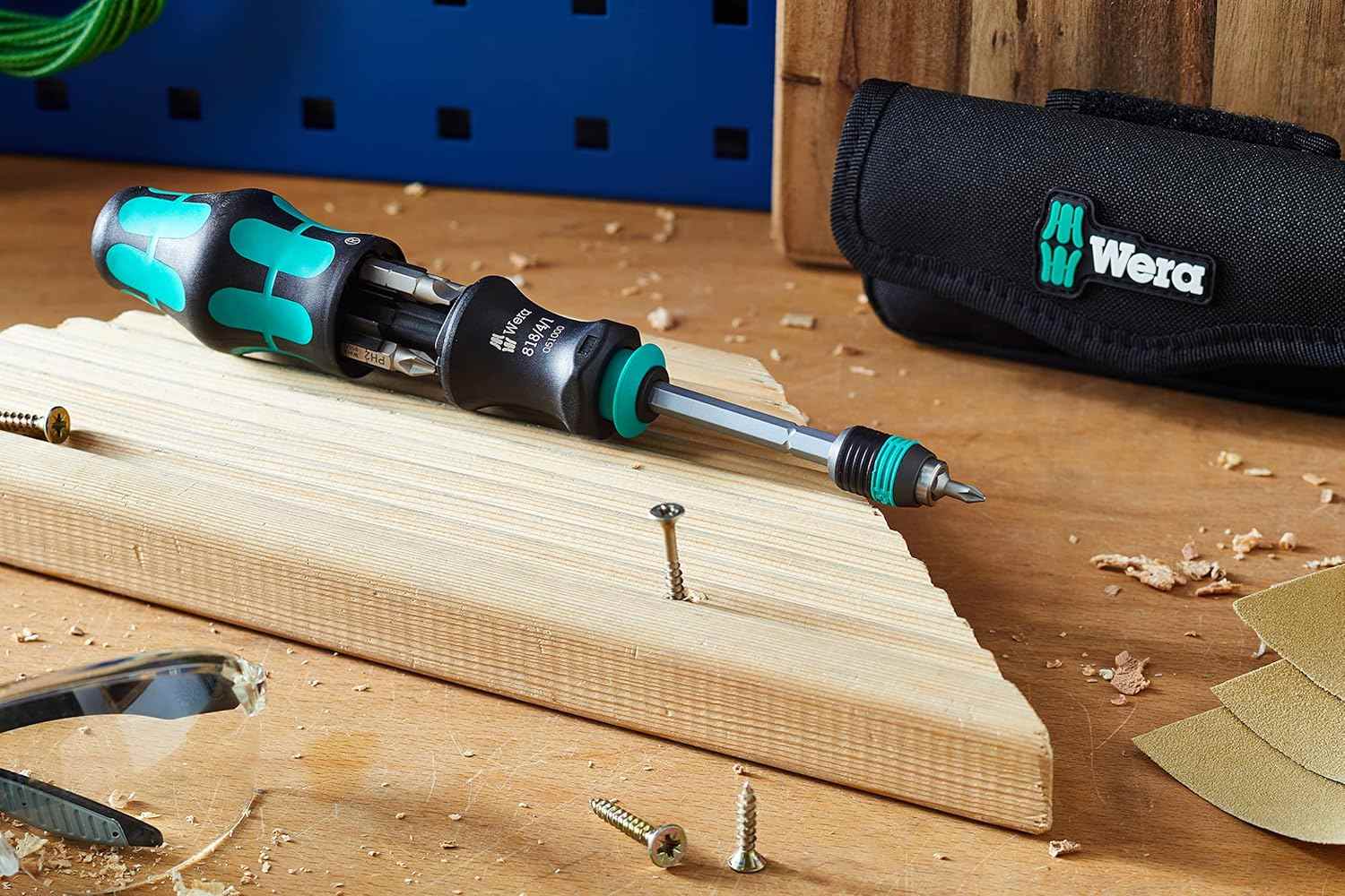 A lifestyle image of the Wera 05051021001 set on a wooden workbench, showcasing the screwdriver with the bit magazine open alongside the textile storage pouch and various wood screws.