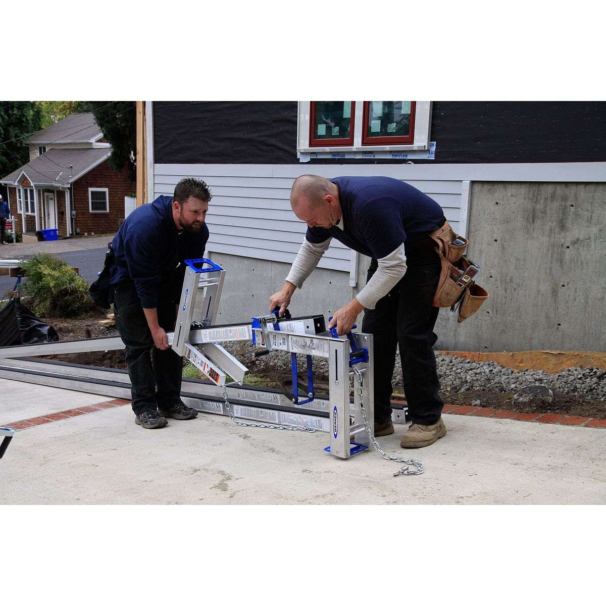 Two construction workers on a driveway preparing the scaffolding system by installing the pump jack mechanism onto the base of the pole.