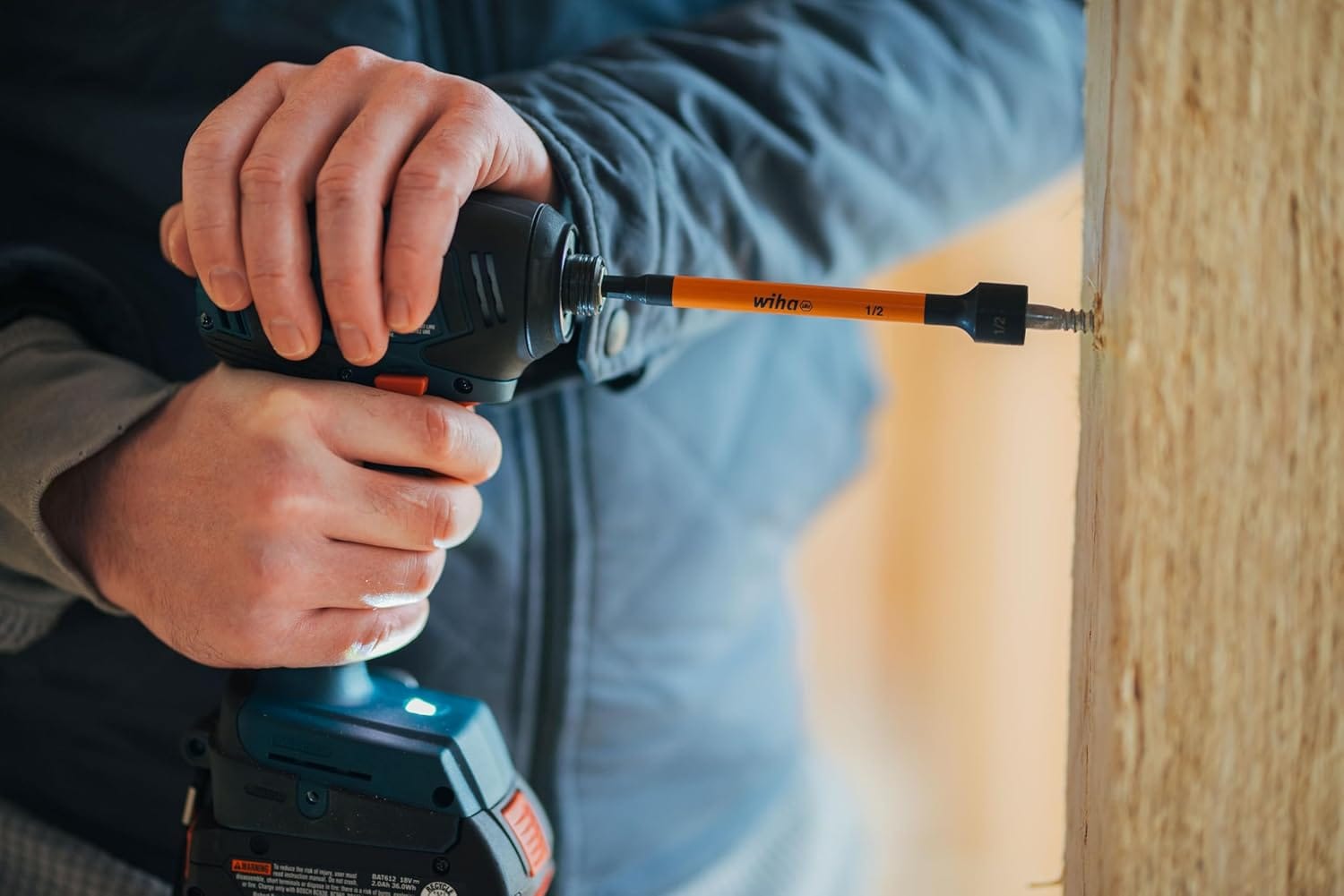 A close-up lifestyle action photo of a tradesman's hands firmly guiding a cordless impact driver to securely drive a hex head screw into a structural wood timber post, utilizing an extended 6-inch orange 1/2-inch Wiha magnetic nut setter socket bit.