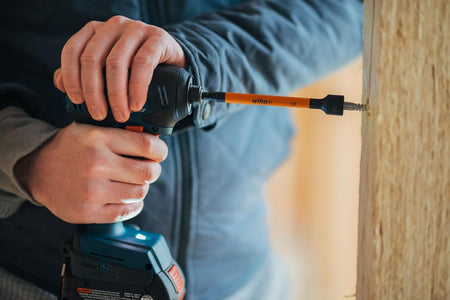 A close-up lifestyle action photo of a tradesman's hands firmly guiding a cordless impact driver to securely drive a hex head screw into a structural wood timber post, utilizing an extended 6-inch orange 1/2-inch Wiha magnetic nut setter socket bit.