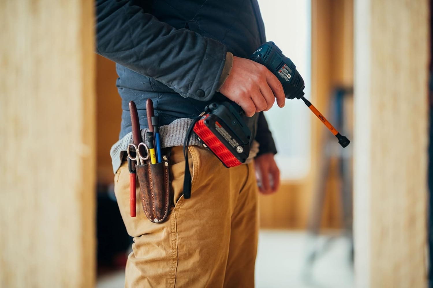 A close-up application photo focusing on a professional technician on a construction jobsite wearing brown canvas pants and a dark jacket, featuring an attached leather tool pouch carrying trade tools and a cordless power impact driver fitted with an extended 6-inch blue Wiha magnetic nut setter bit.