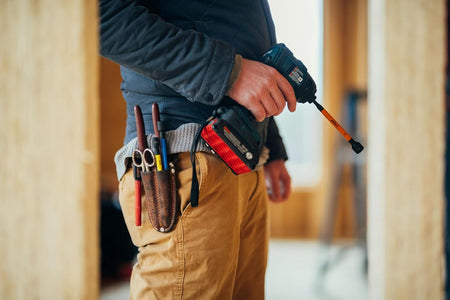 A close-up application photo focusing on a professional technician on a construction jobsite wearing brown canvas pants and a dark jacket, featuring an attached leather tool pouch carrying trade tools and a cordless power impact driver fitted with an extended 6-inch blue Wiha magnetic nut setter bit.