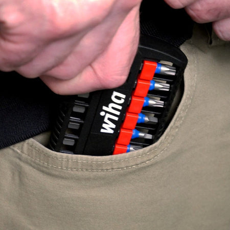 Hand placing a compact Wiha bit set into a beige pants pocket; bits are organized in a black holder with red and blue sections.