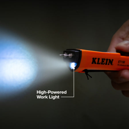A hand holding a Klein ET11M high-powered work light flashlight, which is glowing with a bright blue-white light. The flashlight has an orange body with black accents and the brand name 'KLEIN' printed on it.