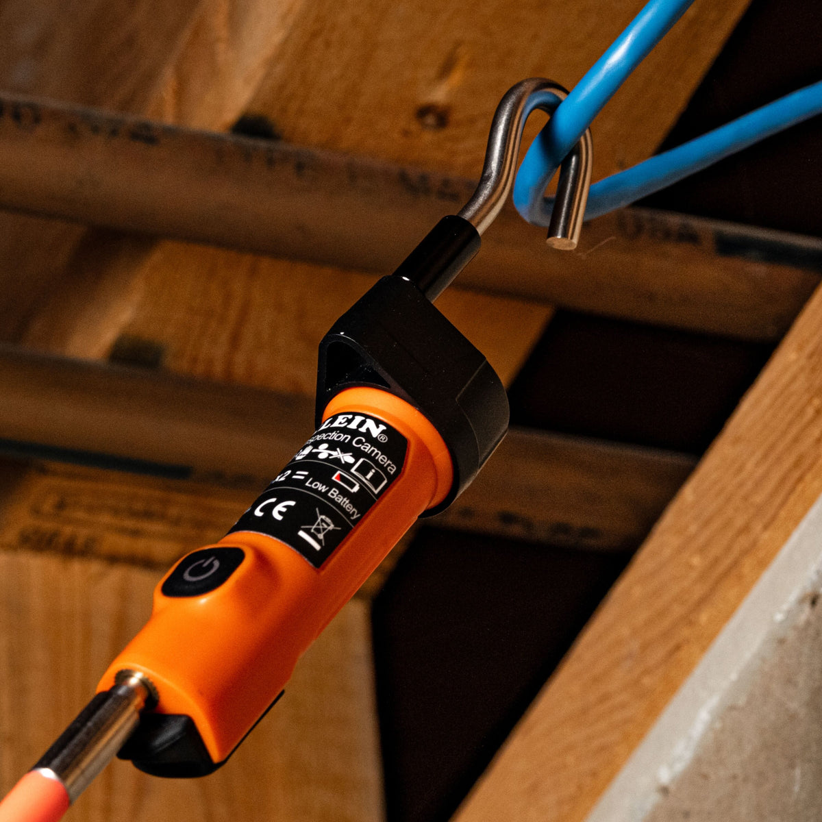 An orange and black BIN brand electric chain saw extension bar with a silver metal hook, shown attached to a blue cable in a wooden attic or workshop environment.