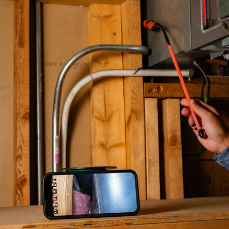 A hand holds an orange laser pointer at an angle above a smartphone screen, which displays a live feed of the laser beam traveling upward through metal and white pipes against a wooden wall.