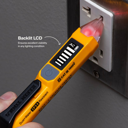 A yellow and black STICKElectric CAT 12 1000V multimeter tester plugged into a wall outlet, with a backlit LCD screen showing a red illuminated indicator. A hand is holding the tool, and text on the image highlights the backlit LCD feature.