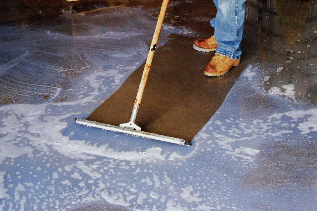 A person in work boots uses a floor squeegee to clean a sudsy concrete floor, demonstrating the BLUE BEAR 700DG degreaser in action.