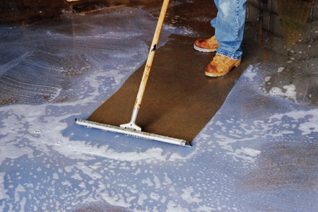 A person in work boots uses a floor squeegee to clean a sudsy concrete floor, demonstrating the BLUE BEAR 700DG degreaser in action.