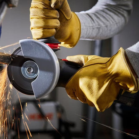 A close-up action shot of an operator in yellow protective gloves using the Bosch 5 inch angle grinder to cut through a metal bar, producing bright sparks.