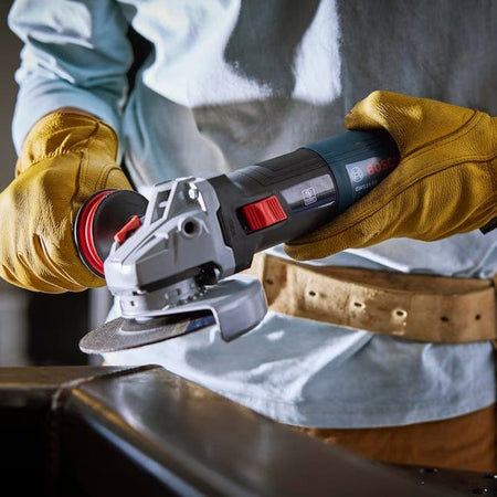 An action view of an operator using the corded Bosch angle grinder to surface a thick metal plate on a workbench, demonstrating the use of the side handle for stability.