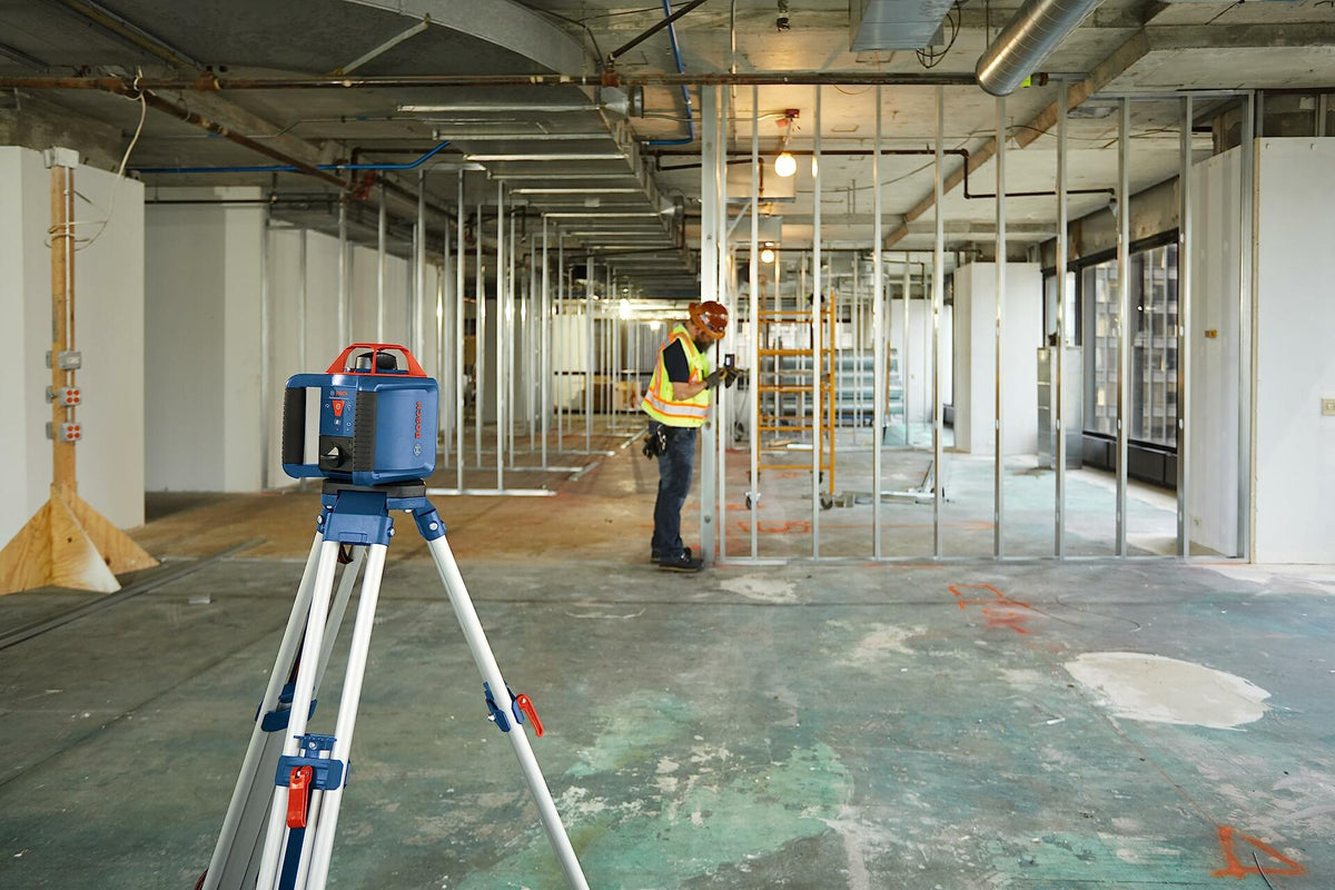 Jobsite action shot showing a Bosch rotary laser mounted on a tripod in the foreground, projecting a level plane, while a construction worker is seen in the background installing metal wall studs
