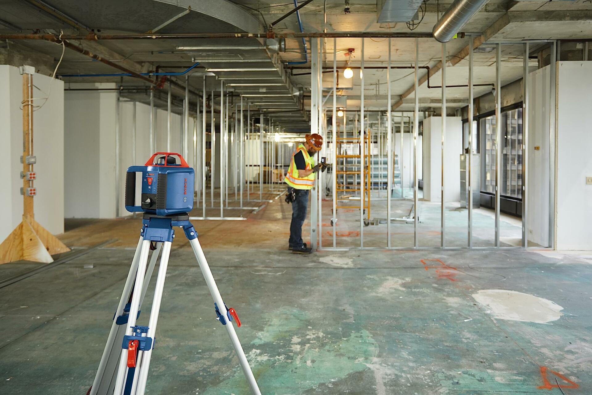 Jobsite action shot showing a Bosch rotary laser mounted on a tripod in the foreground, projecting a level plane, while a construction worker is seen in the background installing metal wall studs