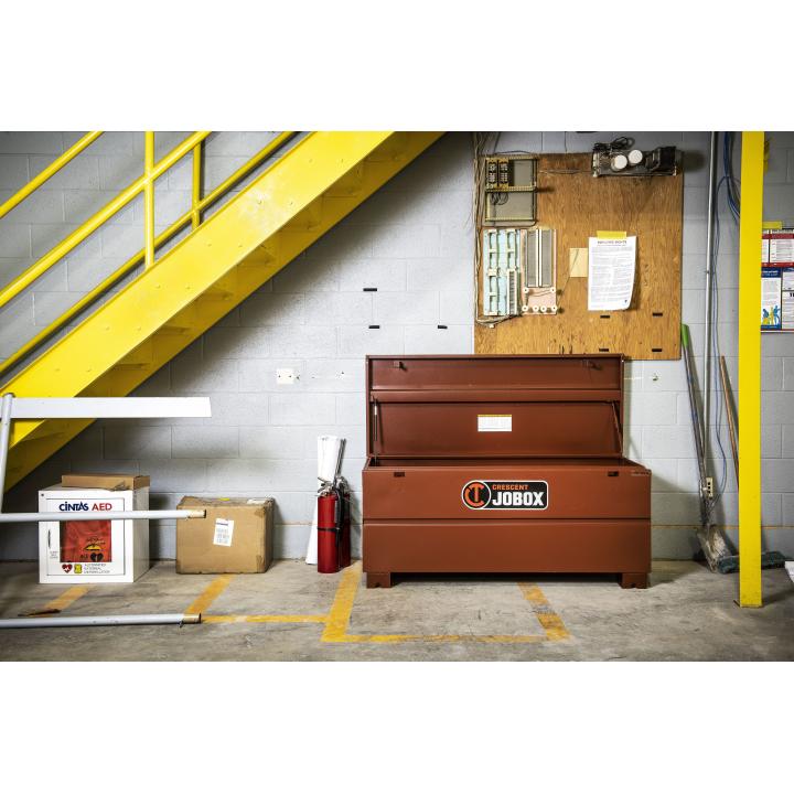 JOBOX 60" Tradesman Steel Chest open. This is a wide shot of a brown rectangular tool chest with an open lid, positioned in a room with a yellow staircase and various equipment against a white brick wall. The chest has a black and orange logo that says "Crescent JOBOX" and is surrounded by a fire extinguisher, a first aid kit, and cardboard boxes.